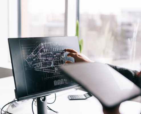 A person points at architectural blueprints displayed on a computer monitor while holding a tablet, working in a bright office with large windows in the background.