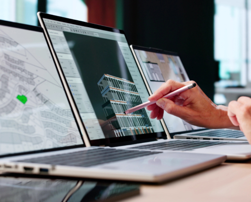 A person points at a 3D building model on a laptop screen with a pen. Two laptops beside it display architectural plans and diagrams. The workspace is modern and professional.