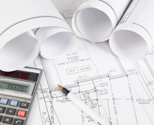 Rolled and flat architectural blueprints with technical drawings, a calculator displaying “96600,” and a black-and-white pen, all arranged on a clean white desk.