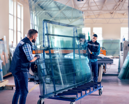 Two workers in a factory move a large glass sheet using a wheeled cart. They are wearing casual work clothes and gloves. The workspace is bright with high ceilings, large windows, and stacks of glass in the background.