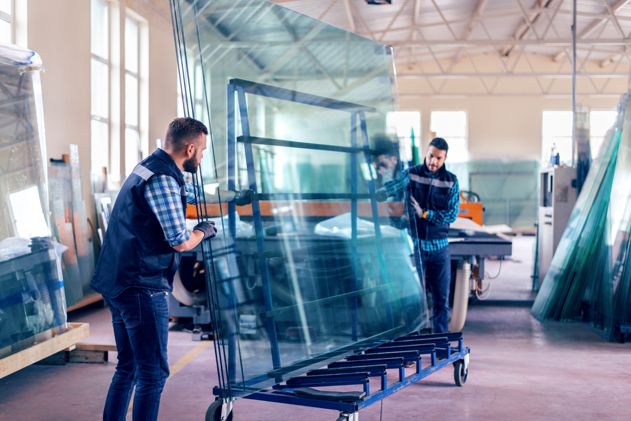 Two workers in a factory move a large glass sheet using a wheeled cart. They are wearing casual work clothes and gloves. The workspace is bright with high ceilings, large windows, and stacks of glass in the background.