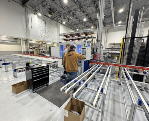 Two people operate a machine in an industrial facility with metal conveyor rails, a black tool cart, storage shelves, and high ceilings. Boxes and equipment are visible throughout the well-lit workspace.