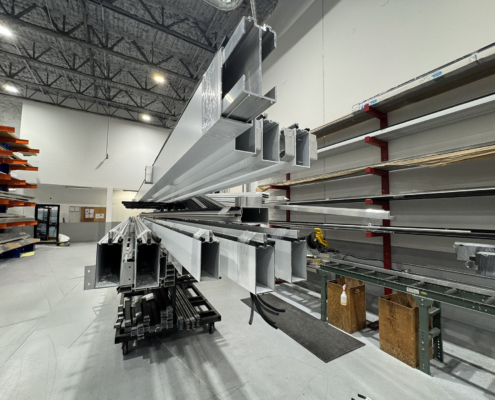 Long aluminum profiles are stacked and stored on metal racks inside a spacious warehouse with high ceilings, exposed beams, and organized shelving along the walls.