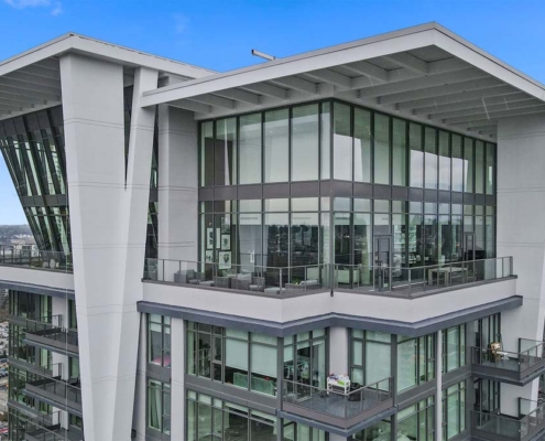 A modern high-rise apartment building with large floor-to-ceiling windows and spacious balconies, set against a clear blue sky and a cityscape in the background.