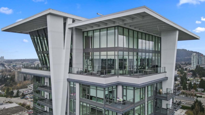A modern high-rise apartment building with large floor-to-ceiling windows and spacious balconies, set against a clear blue sky and a cityscape in the background.