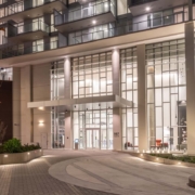 The image shows the well-lit entrance of a modern high-rise building at night, featuring large glass windows, tall white columns, and a circular driveway with landscaped areas and a yellow fire hydrant.