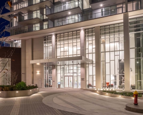 The image shows the well-lit entrance of a modern high-rise building at night, featuring large glass windows, tall white columns, and a circular driveway with landscaped areas and a yellow fire hydrant.