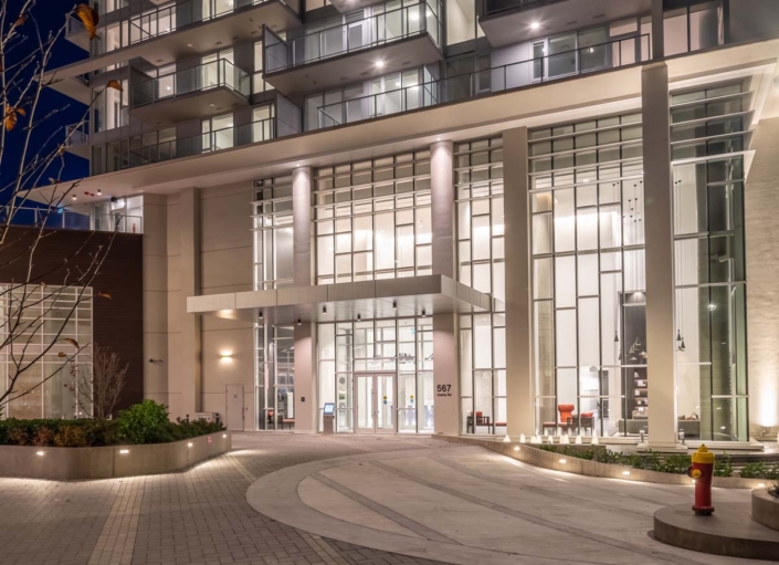 The image shows the well-lit entrance of a modern high-rise building at night, featuring large glass windows, tall white columns, and a circular driveway with landscaped areas and a yellow fire hydrant.