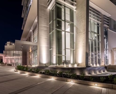 Modern building exterior at night with tall glass windows, illuminated columns, and landscaped pathways. Soft lights highlight architectural details and guide along the concrete path.