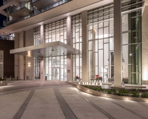Modern building entrance at night with tall glass windows, illuminated columns, well-lit facade, and a landscaped walkway leading to the main doors.