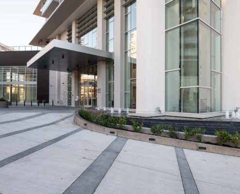 Modern building entrance with large glass windows, tall columns, and a shaded overhang. Curved landscaping with small bushes and a linear fountain feature are visible in the foreground along a broad concrete walkway.