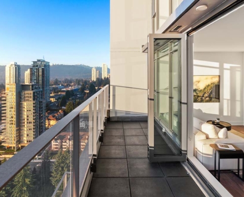 A modern high-rise apartment with an open glass door leads to a balcony overlooking a cityscape with tall buildings, roads, and trees under a clear blue sky.
