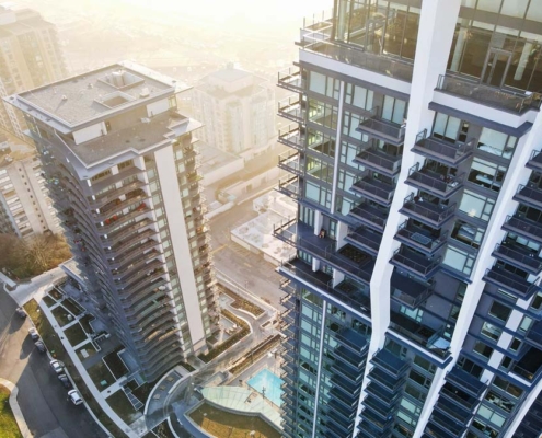Aerial view of two modern high-rise apartment buildings with balconies, surrounded by city streets, smaller buildings, and a pool area below, bathed in warm sunlight.