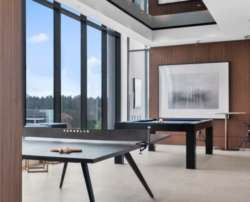 A modern game room with floor-to-ceiling windows, a ping pong table in the foreground, a pool table in the background, wood-paneled walls, and minimalist decor with framed art on the wall.