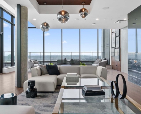 Modern living room with large windows, white sectional sofas, glass coffee table, metallic pendant lights, gray rug, and a marble fireplace. The room has wood floors and panoramic city views through the floor-to-ceiling glass.