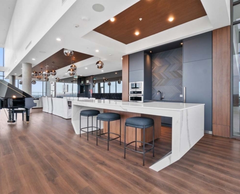 Modern open-concept kitchen and lounge with a marble island, three bar stools, sleek appliances, wooden flooring, pendant lights, and a grand piano by floor-to-ceiling windows with city views.
