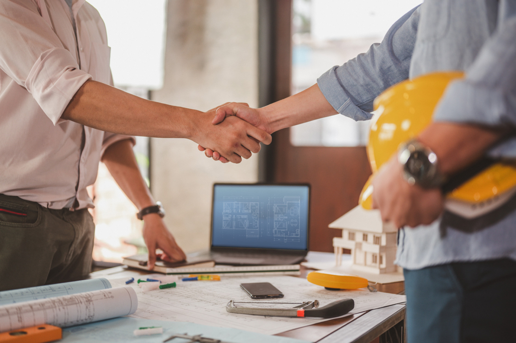 Two people shaking hands over a table with blueprints, a model house, and a laptop; a third person holds a yellow hard hat, suggesting a meeting or agreement in a construction or architectural setting.