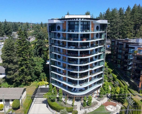 A modern mid-rise apartment building with curved balconies, surrounded by trees and smaller homes, stands in a residential neighborhood under a clear blue sky.