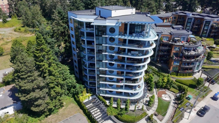 A modern, multi-story residential building with curved balconies and large windows, surrounded by trees and landscaped gardens, viewed from above on a sunny day.