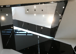 View looking up at a modern interior staircase with black glass railings, angular lines, and hanging ceiling lights. Bright natural and artificial light illuminate the minimalist architecture.