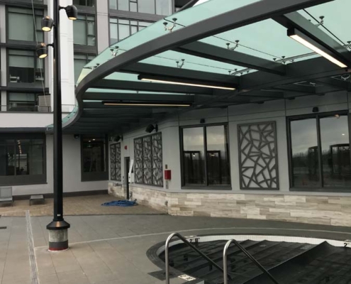 A modern building entrance with a glass canopy, geometric wall panels, a recessed hot tub covered with a black tarp, and a tall pole with multiple lights on a paved patio area.