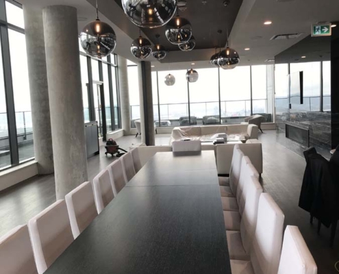 Modern dining area with a long black table, white chairs, large windows, and multiple round pendant lights. The space is bright with natural light and features a city view.