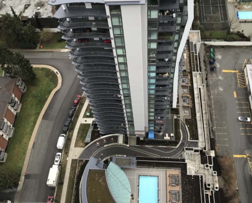 A tall apartment building viewed from above, with balconies facing outward. At the base, there is a rectangular outdoor swimming pool surrounded by lounge chairs and walkways, with nearby roads and greenery visible.