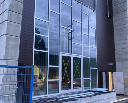 A building under construction featuring tall, narrow glass windows with metal frames, partially unfinished concrete, and construction materials and fencing in the foreground.