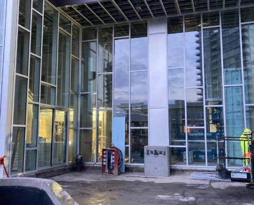 A modern building under construction with large glass windows, exposed ceiling framework, and scattered construction materials. Two workers in safety gear stand near the entrance.