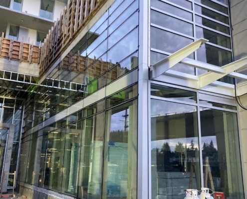 A modern building under construction features large glass windows and metal framing. Construction materials, wooden supports, and gas tanks are visible near the unfinished structure.