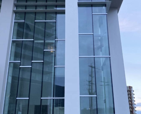 A tall building facade with large glass windows and white vertical structural columns, reflecting the sky and nearby buildings. Some construction materials are visible at the top of the structure.