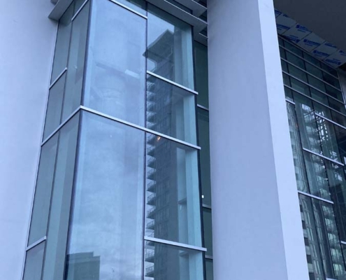 A close-up view of a modern building facade with large glass windows, vertical white columns, and overhanging horizontal architectural features. Another building is reflected in the glass.