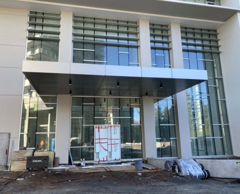 The image shows the entrance of a modern building with large glass windows, a metal canopy, and a door covered with protective material. Construction equipment and materials are scattered around the unfinished ground.