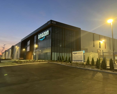 Amazon fulfillment center building at dusk, with illuminated Amazon signage on glass facade, exterior lights on, and a sign reading Amazon Fulfillment near the entrance. The sky is dimly lit, and the parking lot appears empty.