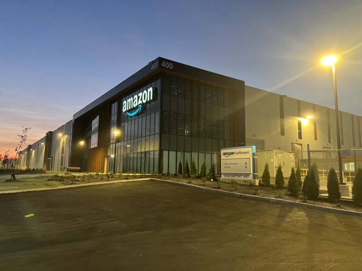 Amazon fulfillment center building at dusk, with illuminated Amazon signage on glass facade, exterior lights on, and a sign reading Amazon Fulfillment near the entrance. The sky is dimly lit, and the parking lot appears empty.