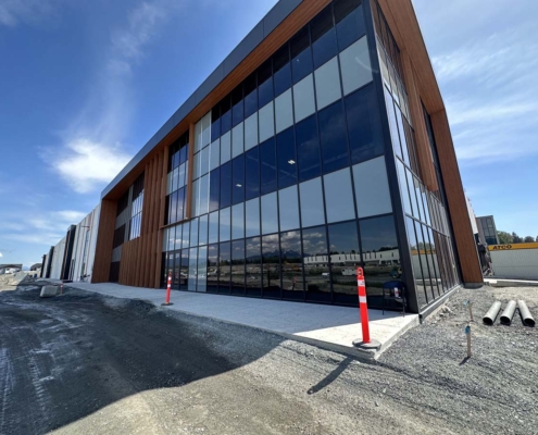 Modern commercial building with large glass windows and wood panel accents, under construction. There are construction cones, dirt, and construction materials around the site, with a partly cloudy sky above.