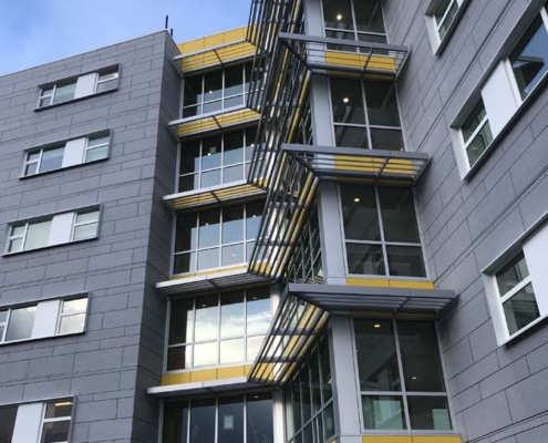 A modern multi-story building with gray exterior panels, large windows, and yellow accents on each floor. Glass awnings cover the entrance, and metal railings frame the balconies. The sky is clear and blue.