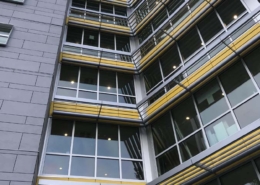 A modern multi-story building with large windows, yellow accents, and horizontal metal shades, viewed from below. The sky is clear, and the building features glass panels at the entrance.