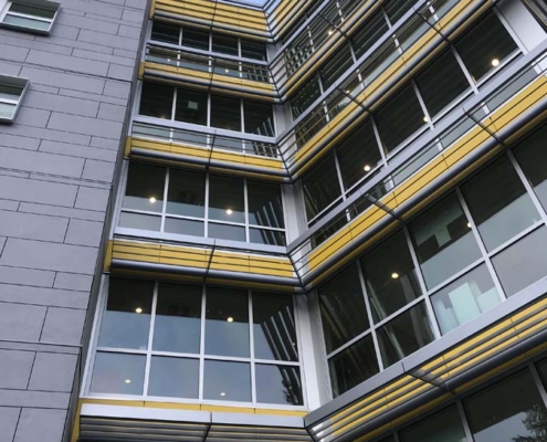 A modern multi-story building with large windows, yellow accents, and horizontal metal shades, viewed from below. The sky is clear, and the building features glass panels at the entrance.