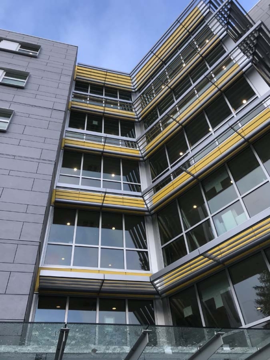 A modern multi-story building with large windows, yellow accents, and horizontal metal shades, viewed from below. The sky is clear, and the building features glass panels at the entrance.