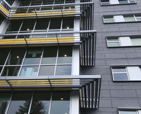 A modern building facade with large windows, yellow horizontal accents, and metal sunshades under a blue sky. The exterior is gray with geometric lines and multiple stories visible.