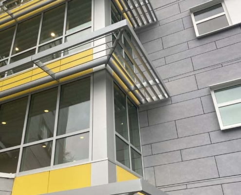 Modern building facade with gray and yellow accents, large glass windows, metal framing, and angled sunshades above each window. The wall surface has horizontal lines and several windows, some slightly open.