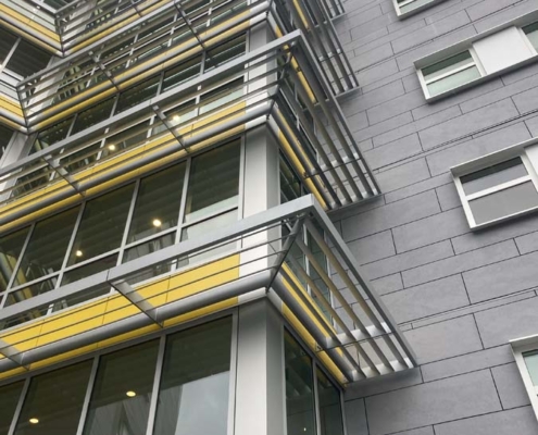 Modern building exterior with large glass windows, gray walls, and yellow accents. Metal sunshades jut out above the windows, creating a geometric pattern. The photo is taken from a low angle, looking upward.