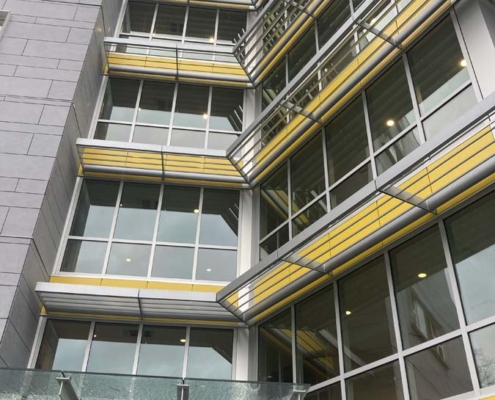 A modern building with large glass windows and horizontal yellow accents, viewed from below. Metal railings and sunshades project outward, and a glass awning covers the entrance at the bottom.