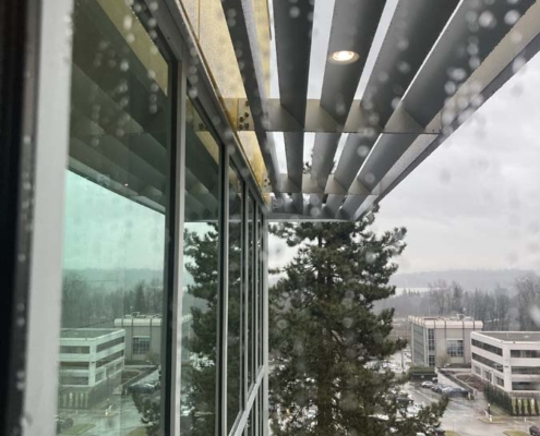 View from a building with glass windows and a slatted overhang, showing raindrops on the glass, pine trees, and office buildings in a parking lot on a cloudy day.