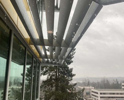 View from a building with metal sunshades and glass windows, looking out over a rainy, cloudy day with trees, parked cars, and office buildings visible in the background.