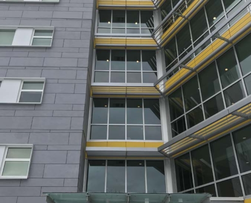 A modern multistory building with gray walls, large windows, and yellow horizontal accents above the windows. There is a glass canopy above the entrance.