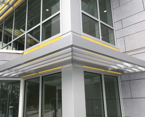 Corner of a modern building with large glass windows, metal and concrete surfaces, and yellow accent lines. Reflections in the windows show trees and the surrounding area.