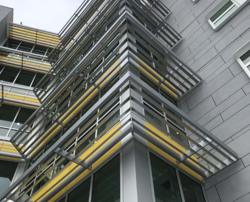 A modern building with large glass windows and silver and yellow metal sunshades, viewed from below against a cloudy sky. The geometric structure creates sharp angles and lines.