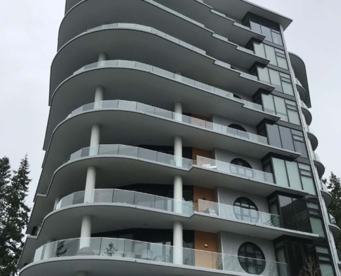 A modern multi-story building with curved balconies, circular cutouts, glass railings, and a unique overhanging roof feature, viewed from below against a cloudy sky. Trees and bare branches are visible at the sides.